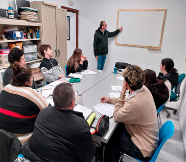 Grupo de personas adultas sentadas alrededor de una mesa en un aula, atendiendo a un hombre que escribe en una pizarra. La clase parece ser de matemáticas o inglés, y los participantes toman apuntes.