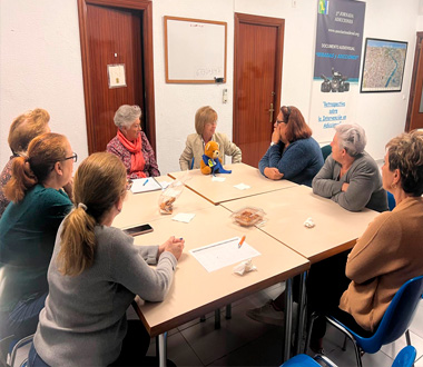 Grupo de mujeres adultas sentadas alrededor de una mesa en una sala, participando en una reunión o taller. Sobre la mesa hay papeles, bolígrafos y algo de comida. El ambiente es acogedor y de conversación.
