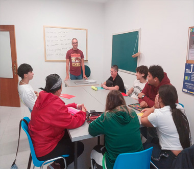 Grupo de jóvenes sentados alrededor de una mesa en un aula, escuchando a un hombre de pie que parece estar impartiendo una clase. En la pared hay una pizarra blanca con texto escrito.