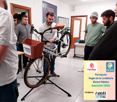 Varios jóvenes observan a un hombre que les enseña a reparar una bicicleta colocada en un soporte. La actividad se realiza en un aula con carteles en la pared. En la esquina inferior derecha aparece un cartel con logos de Cáritas, DESAL y RenovaBike, indicando la colaboración de varias entidades en la actividad.